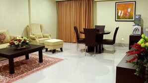Uma sala de estar com sofá, poltrona, mesa de centro e mesa de jantar com cadeiras. Há um quadro na parede e um dispensador de água sobre uma mesa.