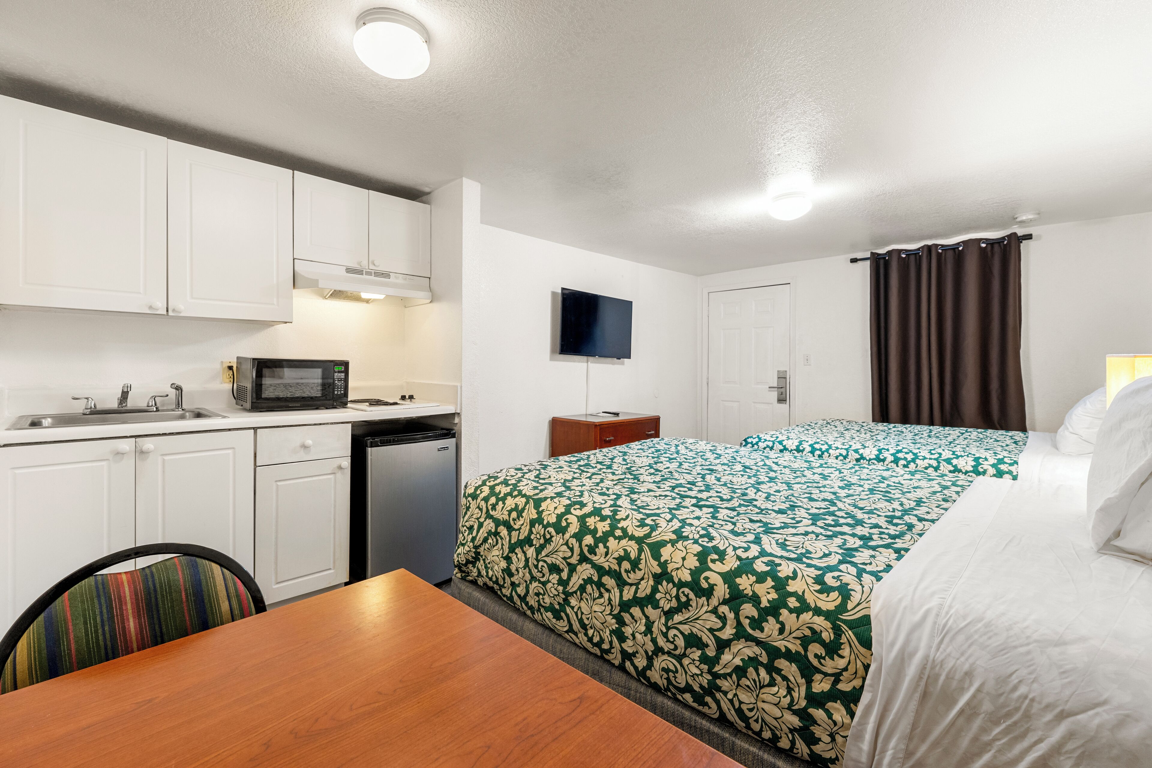 Habitación de hotel compacta con kitchenette, una cama con ropa de cama estampada, televisión y ventana con cortinas.
