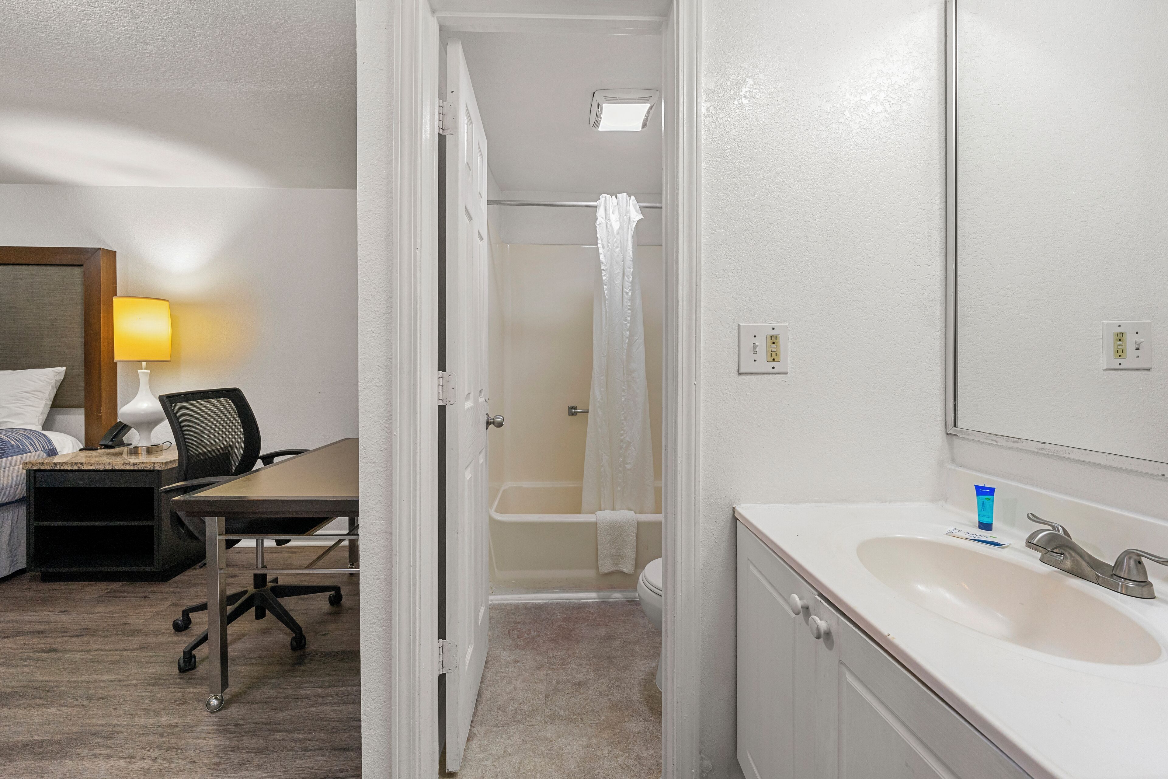 A hotel room with a bed, desk, chair, bathroom with a sink, and a shower.