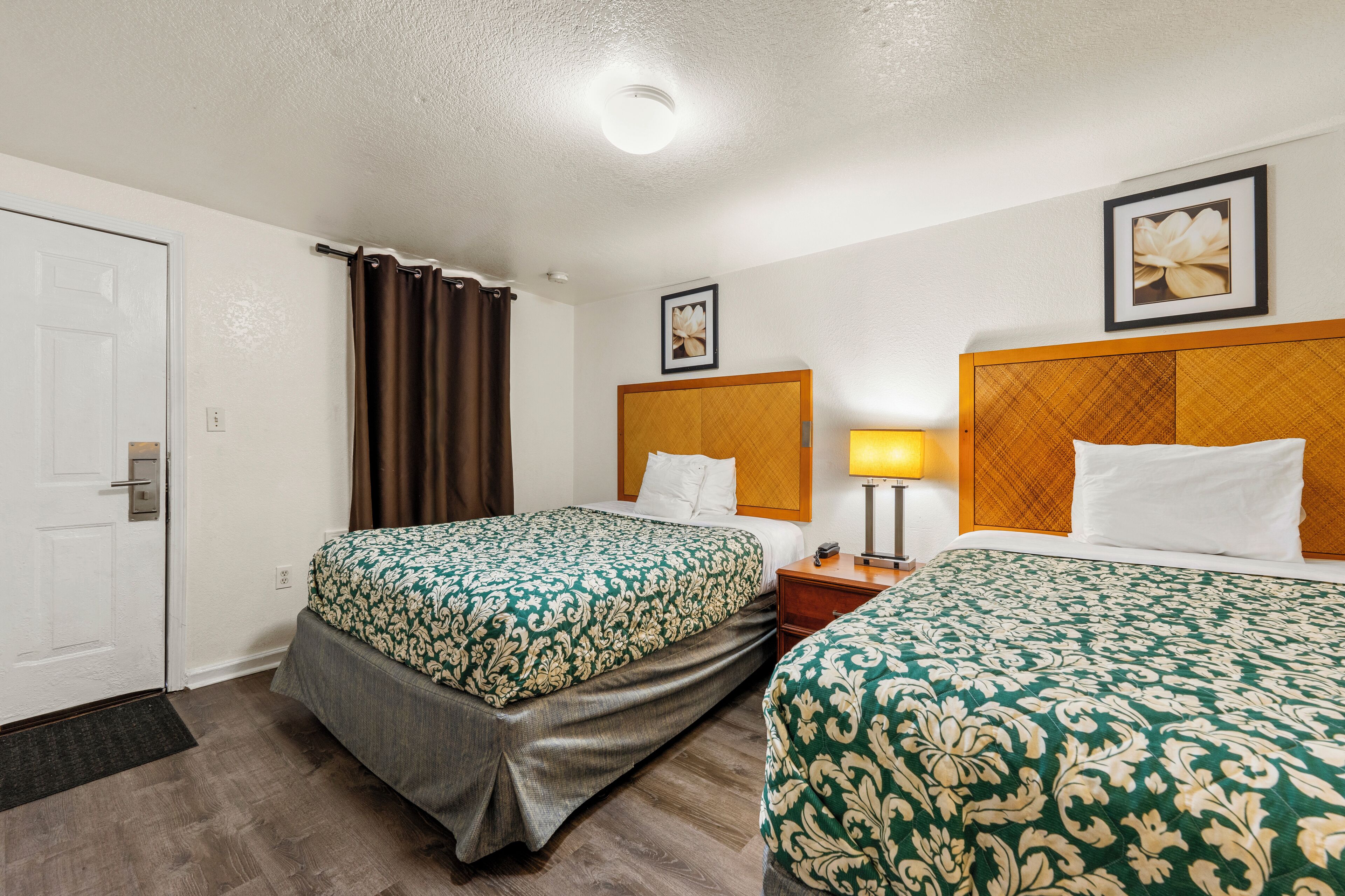 A hotel room with two beds, a wooden headboard, a bedside table with a lamp, and a framed picture on the wall.