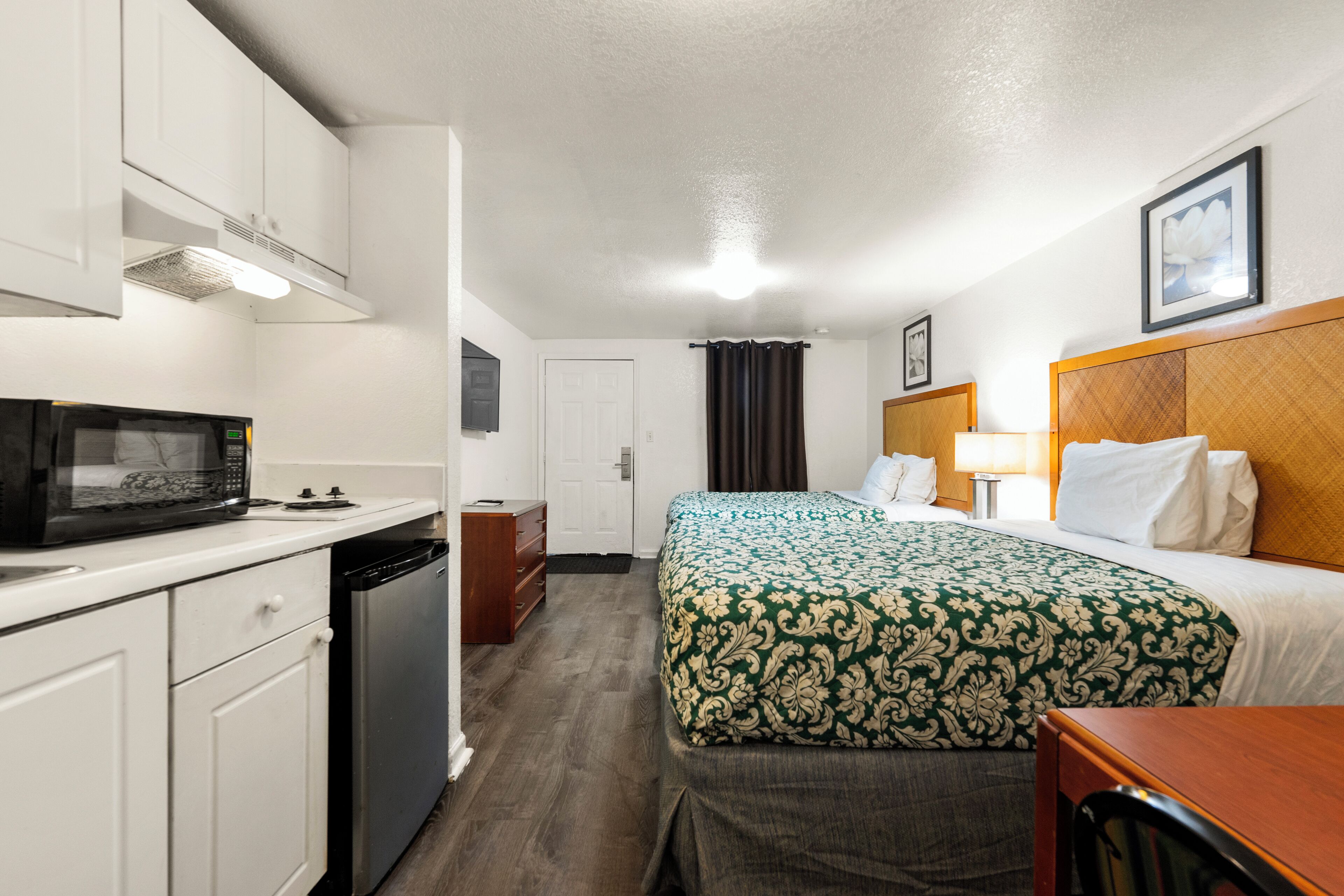 A hotel room with a kitchenette, a bed with a patterned bedspread, a bedside table with a lamp, and a framed picture on the wall.