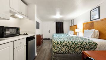 A hotel room with a kitchenette, a bed with a patterned bedspread, a bedside table with a lamp, and a framed picture on the wall.