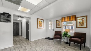 A hotel lobby with a wooden floor, two armchairs, a potted plant, and framed artwork on the walls.