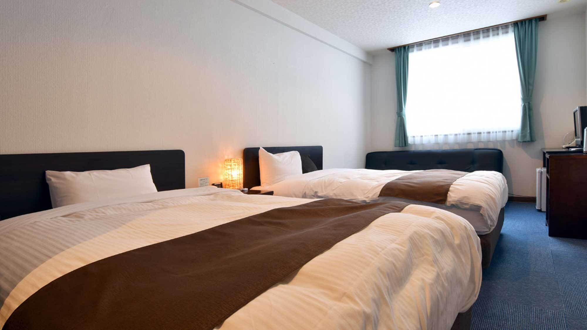 A hotel room with two beds, a nightstand with a lamp, a desk with a computer, and a window with curtains.