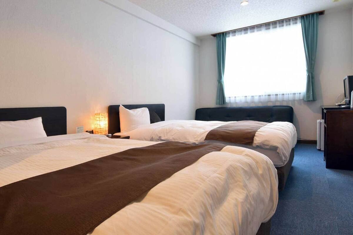 Habitación de hotel con dos camas, un escritorio, una lámpara y un ventanal con cortinas.