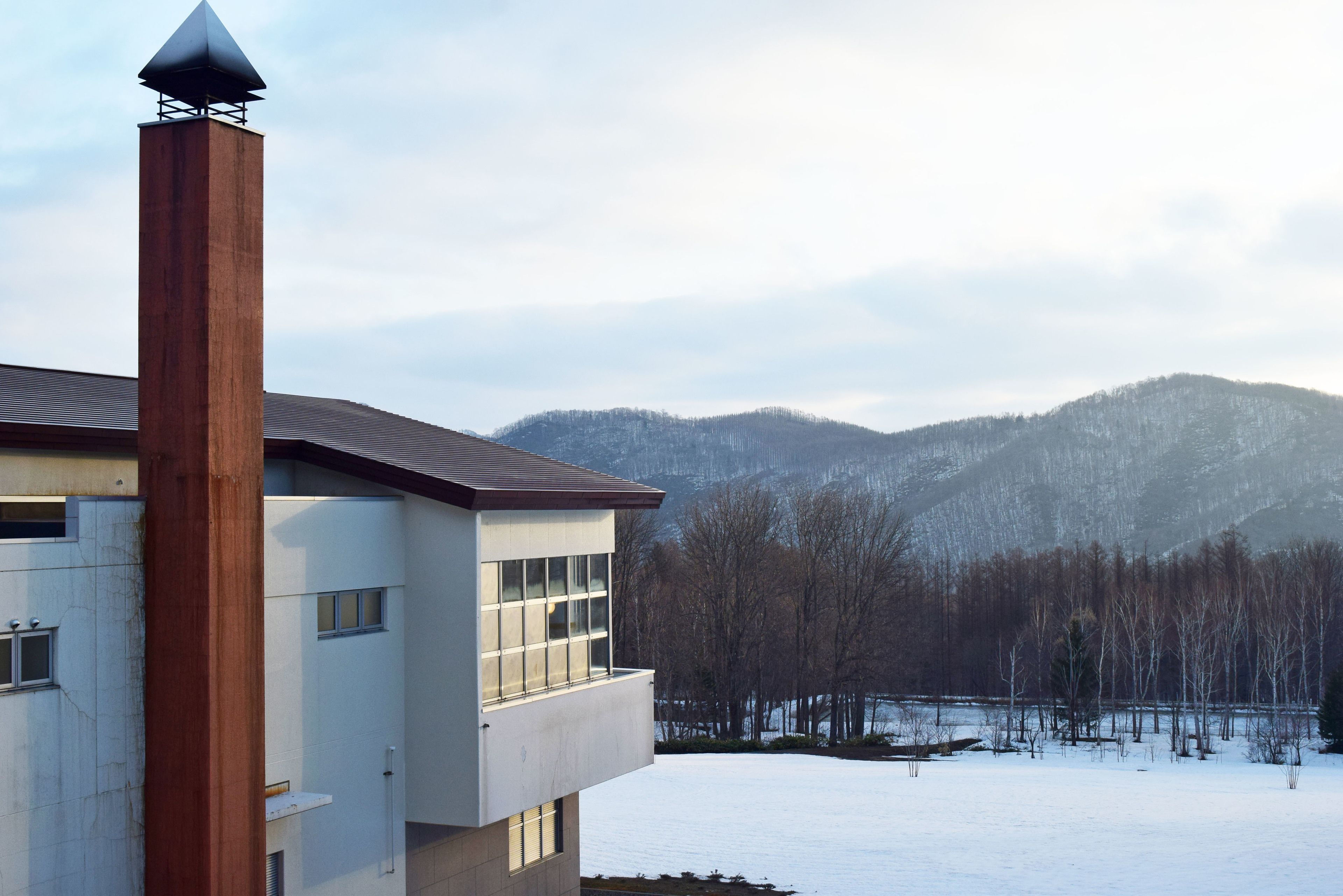 Un edificio moderno con una chimenea destacada, ventanas amplias y vistas a colinas y árboles cubiertos de nieve.