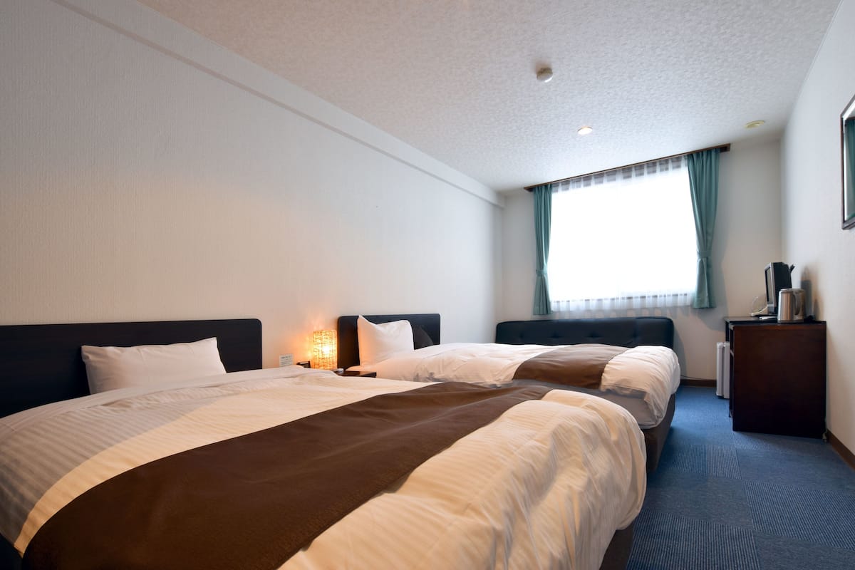 Una habitación de hotel con dos camas, una mesita de noche con lámpara, una ventana con cortinas y un escritorio pequeño con lámpara.