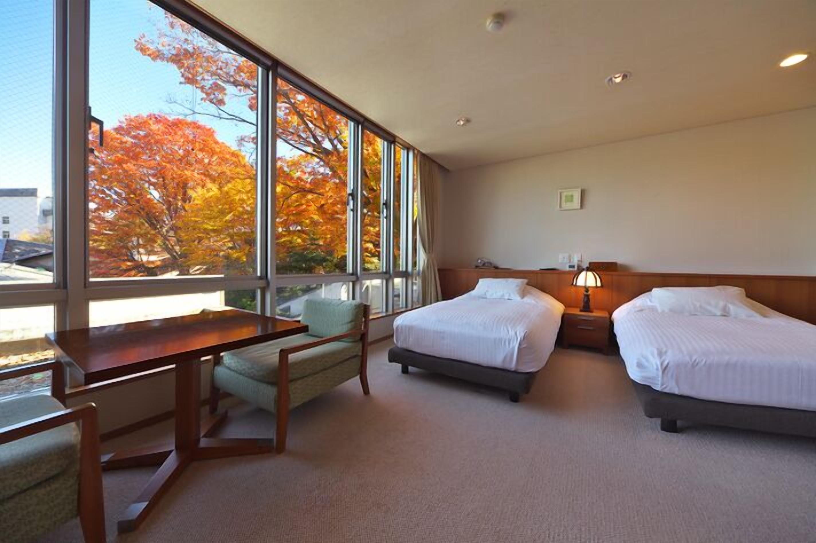 Habitación de hotel con dos camas, un escritorio, una silla y grandes ventanales con vistas a árboles otoñales.