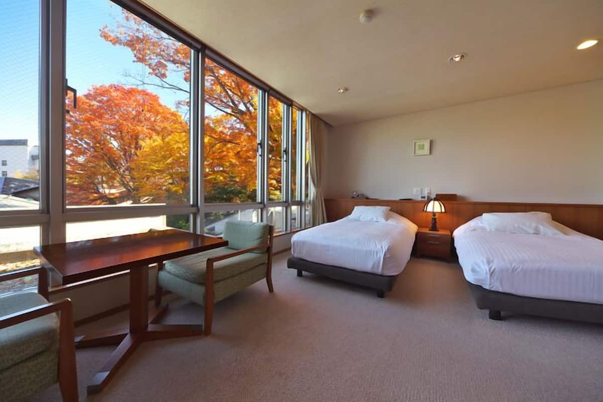 Habitación de hotel con dos camas, un escritorio, una silla y grandes ventanales con vistas a árboles otoñales.