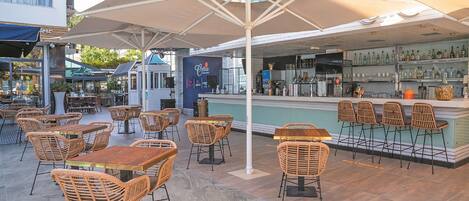 An outdoor seating area with wicker chairs and tables, a bar with a counter, and a large umbrella providing shade.