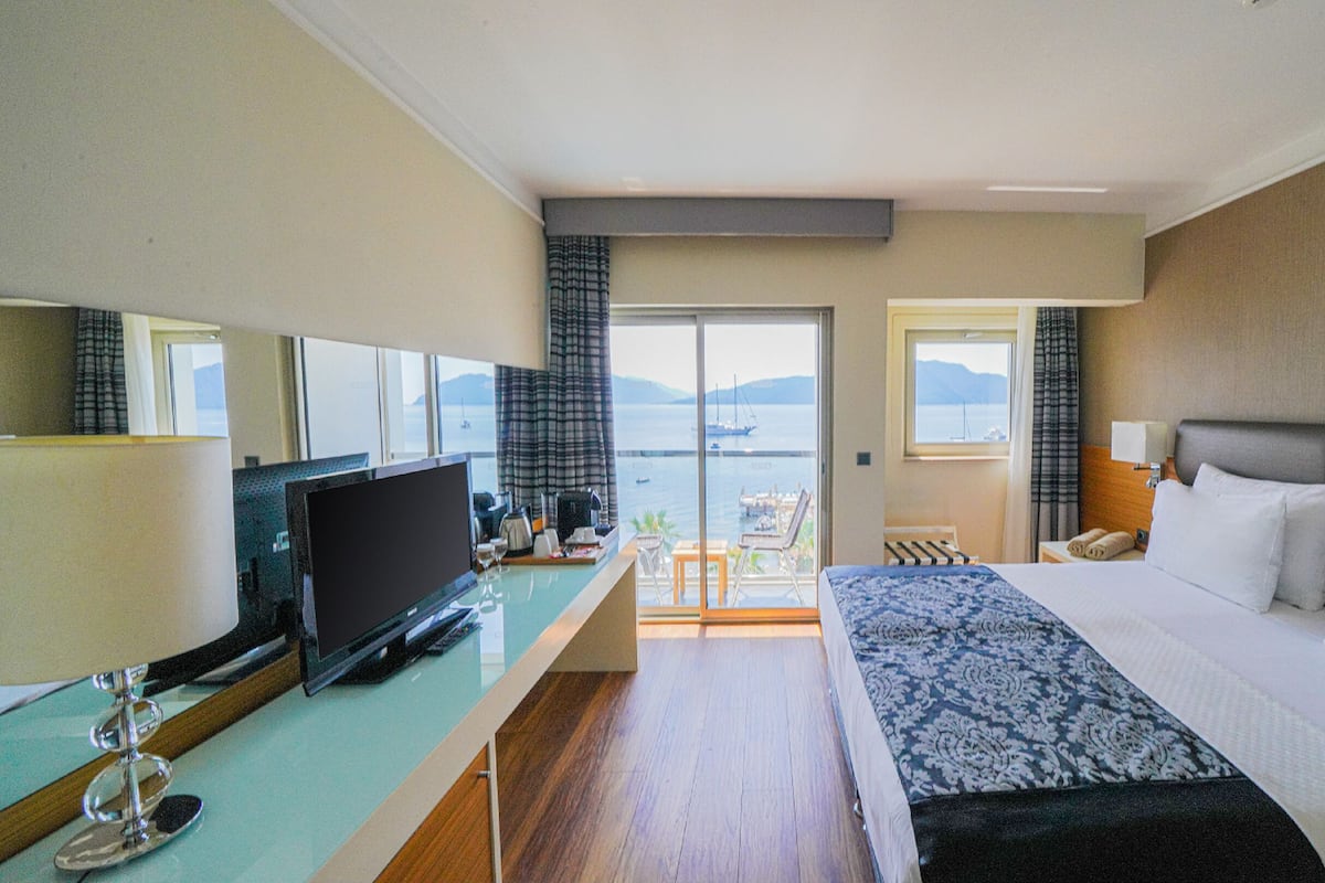 A hotel room with a bed, a desk with a TV, a chair, and a view of the sea.