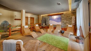 A spa room with wooden flooring, wicker loungers, a green rug, and a stone wall with a mountain painting.