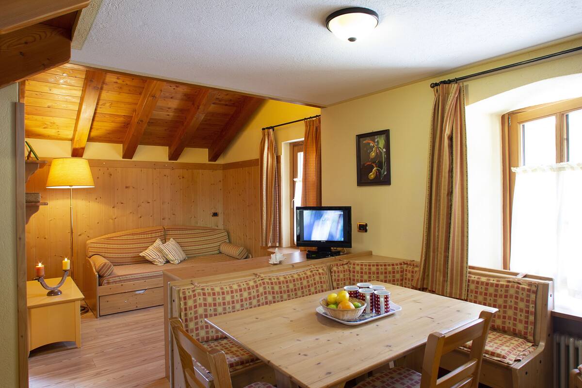 Una habitación acogedora con techo de madera, un sofá, una mesa de comedor con sillas, un televisor y una ventana con cortinas.