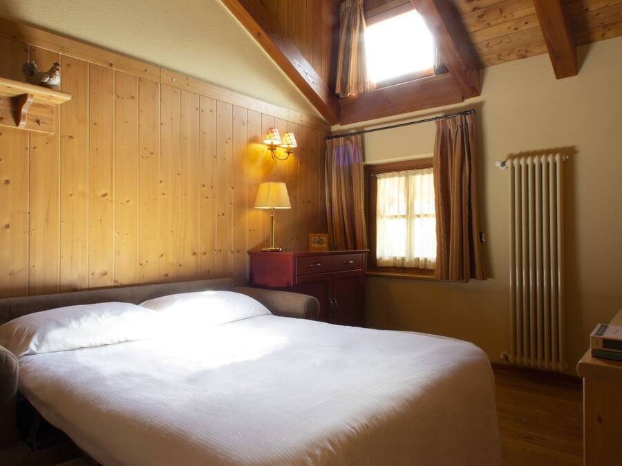 A bedroom with a large bed, wooden walls, a window with curtains, a radiator, and a shelf with a rooster decoration.