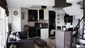 Een moderne woonkamer met een zwarte bank, een bijzettafeltje en een kleine plant. Er is een keukenhoek met zwarte kasten en een betegelde achterwand. Een trap is op de achtergrond zichtbaar.