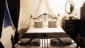 Een slaapkamer met een hemelbed, een nachtkastje met een lamp, een dressoir en een spiegel aan de muur.
