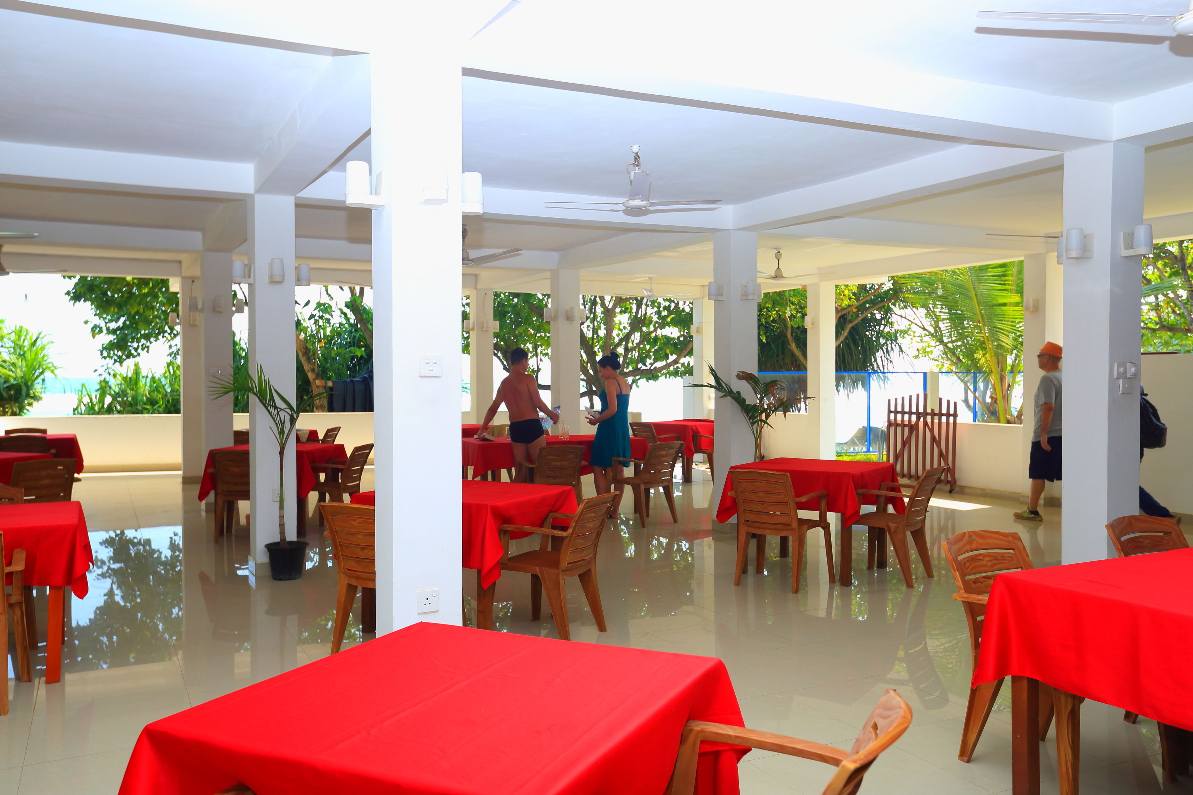 A spacious indoor dining area with red tablecloths and wooden chairs, featuring a high ceiling and large windows with a view of greenery outside.