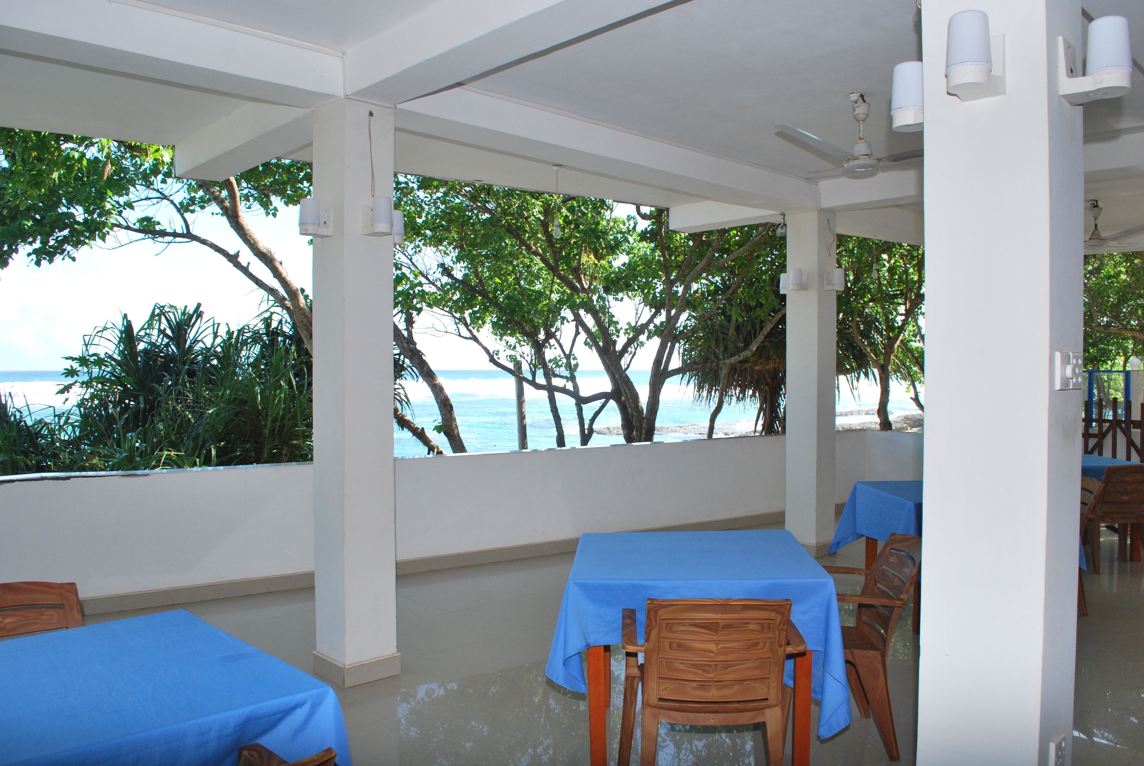 A covered outdoor seating area with tables and chairs, overlooking a beach and trees.