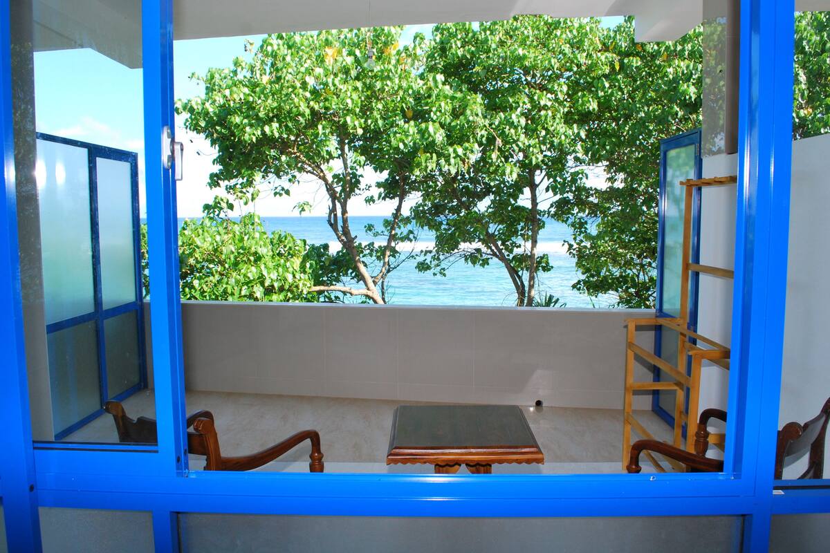 Quarto com vista para o mar e árvores, uma cadeira de madeira, uma mesa pequena e uma janela com moldura azul.