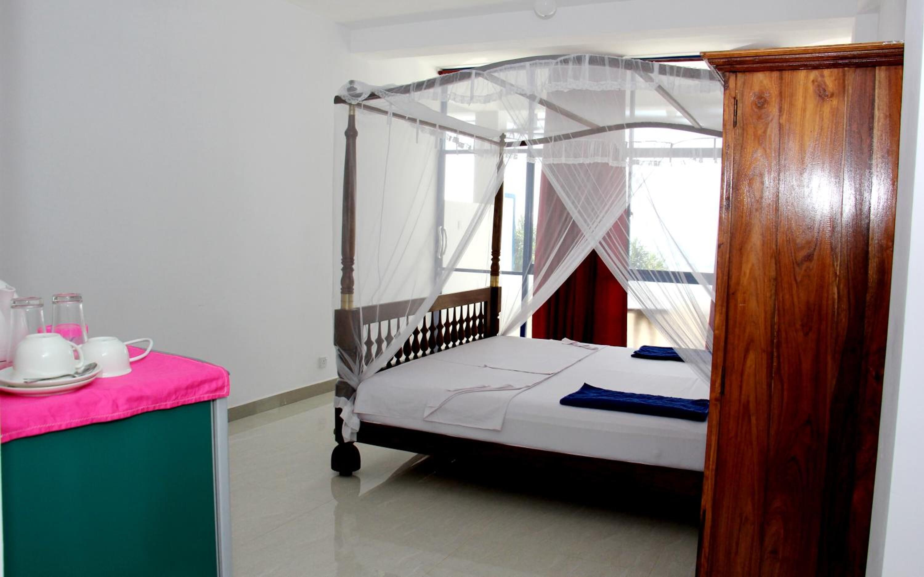 A room with a bed under a canopy, a bedside table with cups and a pink cloth, and a wooden headboard.