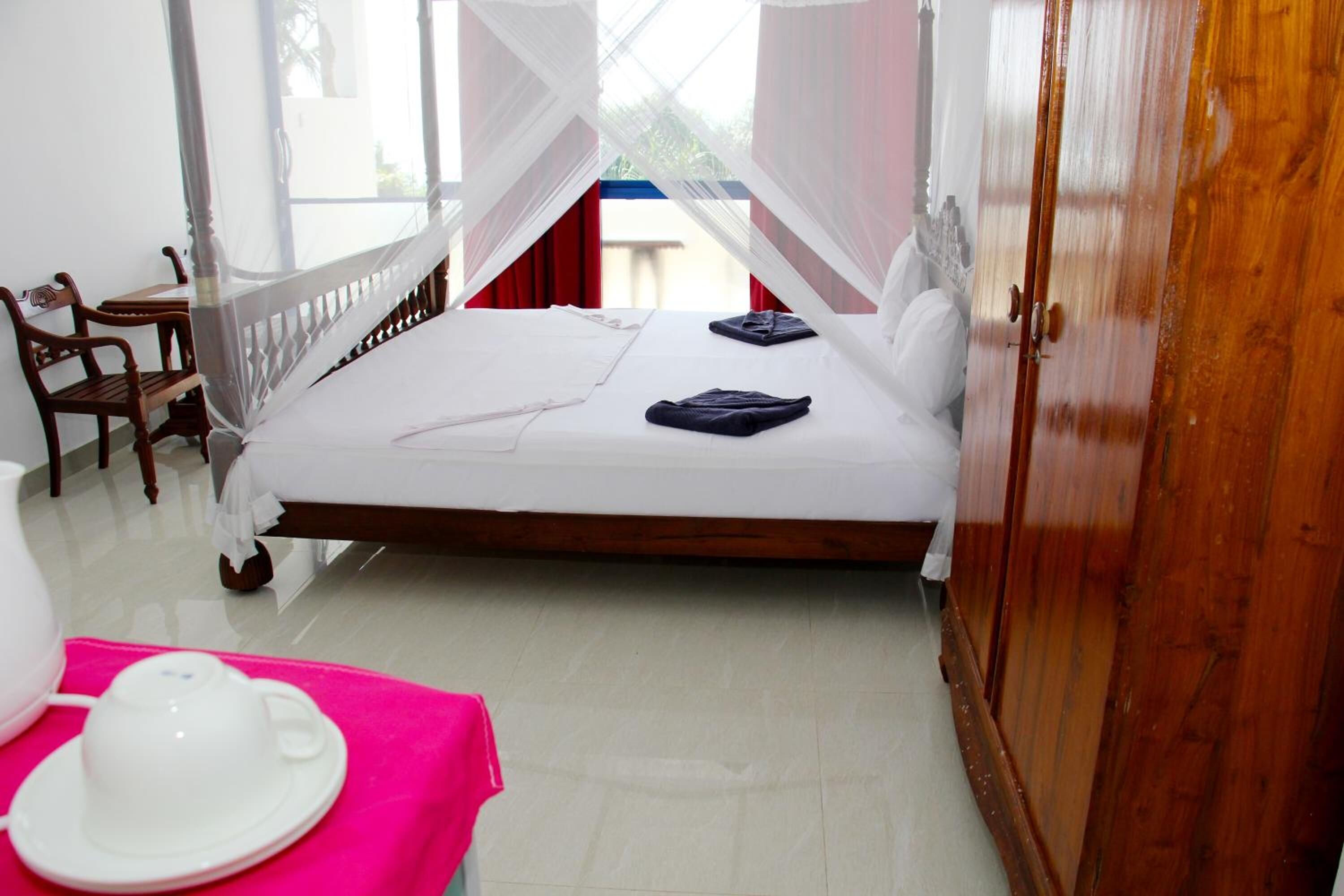 A single bed with a canopy, a wooden wardrobe, a chair, and a table with a pink cloth and a white teapot.