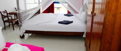 A single bed with a canopy, a wooden wardrobe, a chair, and a table with a pink cloth and a white teapot.