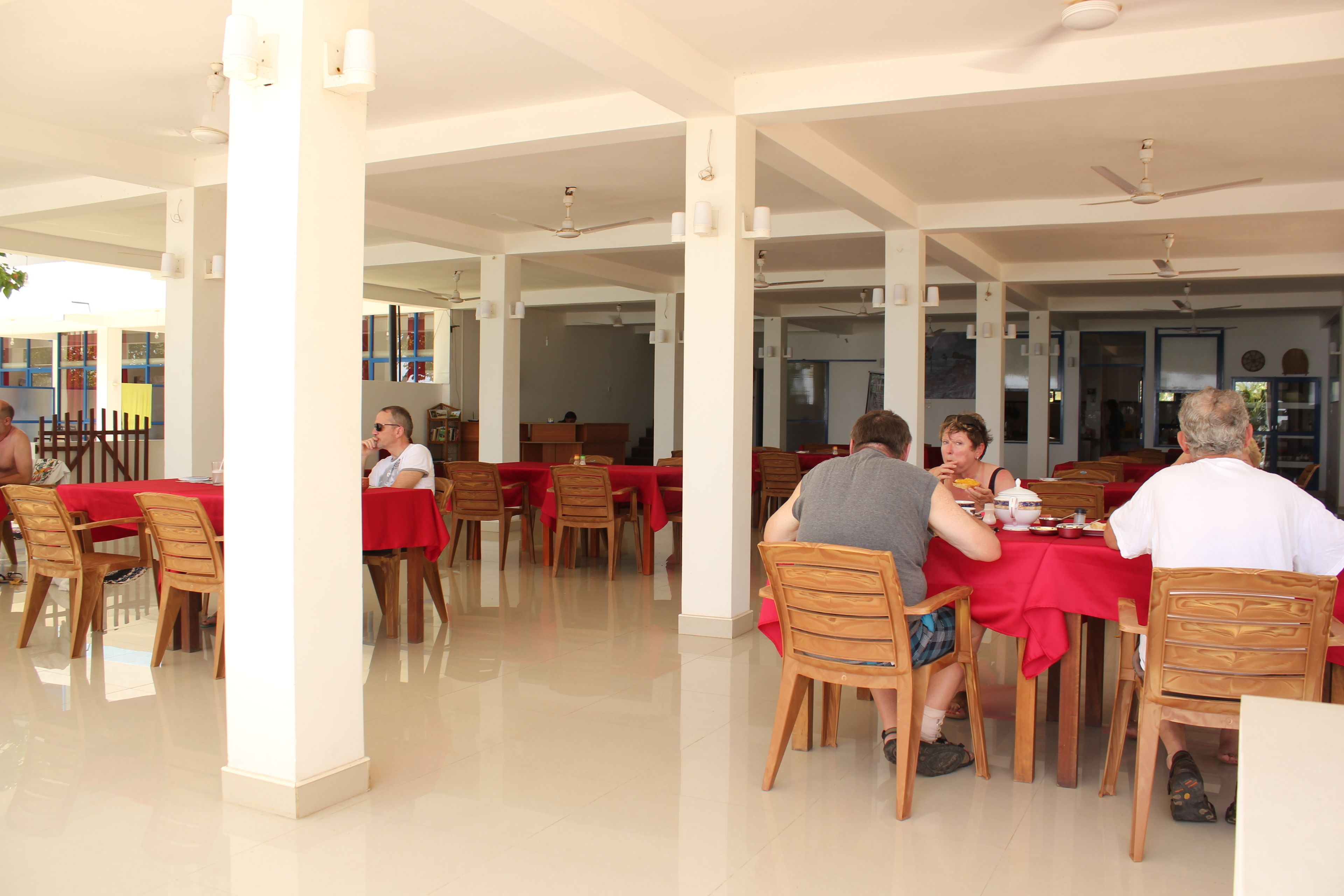 A spacious outdoor dining area with multiple tables and chairs, covered by a roof with ceiling fans.