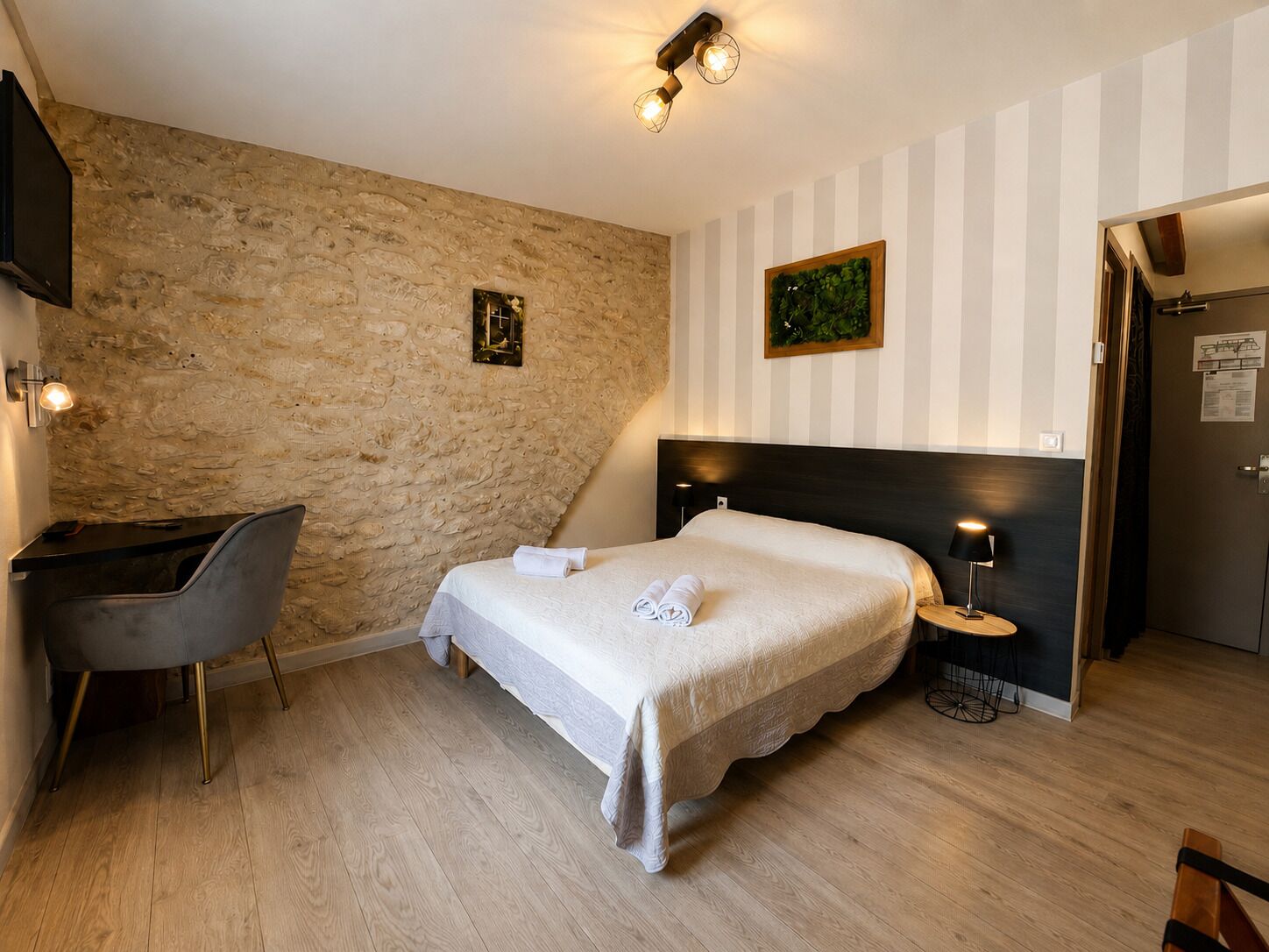 A hotel room with a bed, a desk, a chair, a nightstand, and a door.