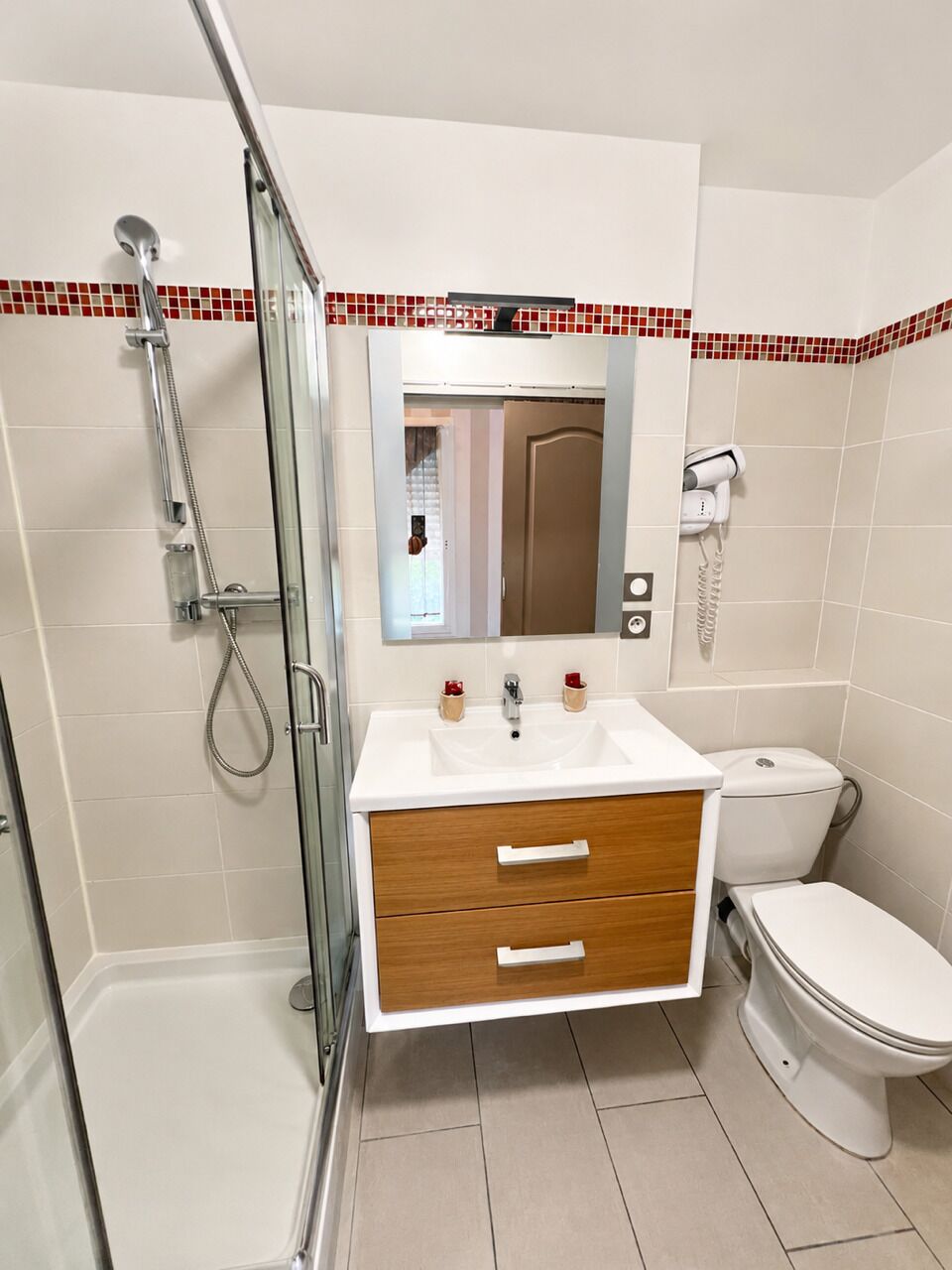 A bathroom with a glass shower enclosure, a white sink with wooden cabinet drawers, a toilet, and a mirror with a red and white tiled border.
