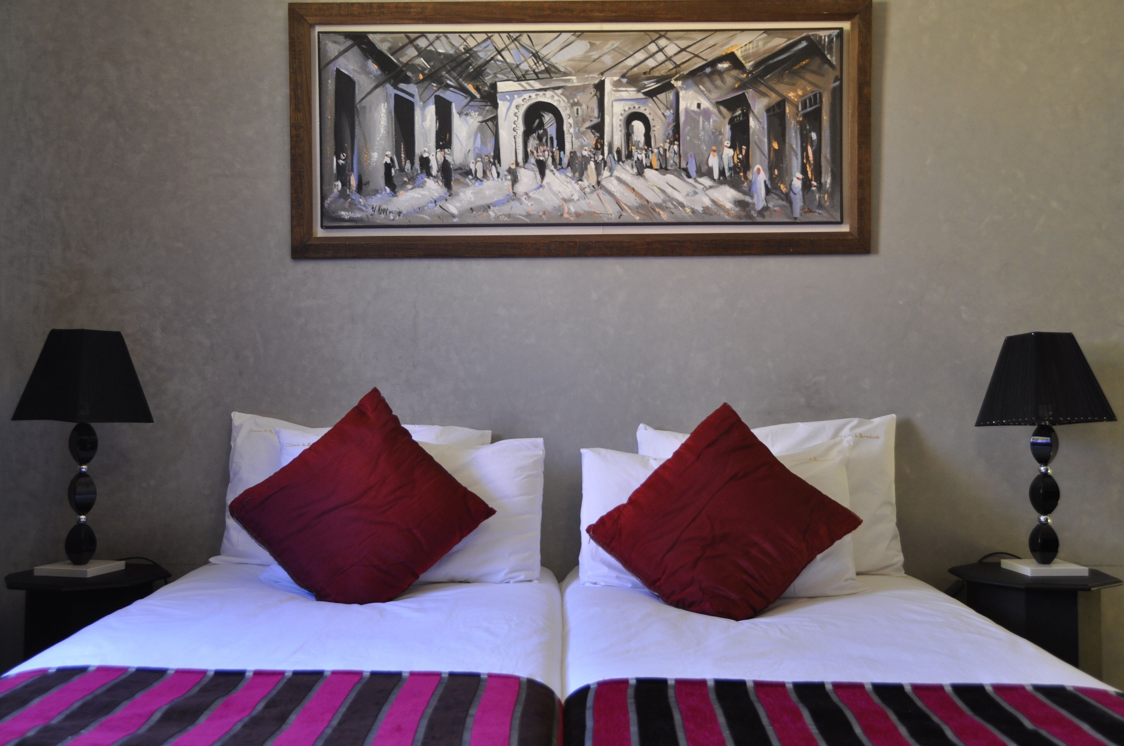 Une chambre d’hôtel avec deux lits, chacun agrémenté de draps blancs et d’oreillers rouges, un tableau encadré au-dessus des lits et deux lampes de chevet.