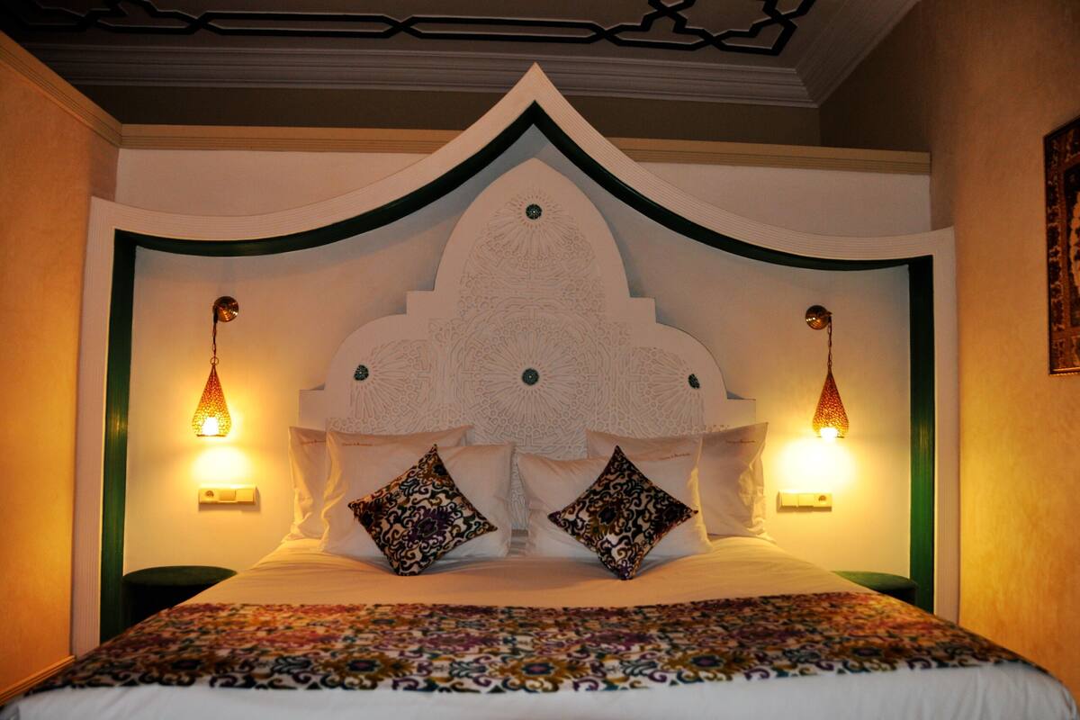A hotel room with a large bed, decorative headboard, two wall-mounted lamps, and a patterned bedspread.