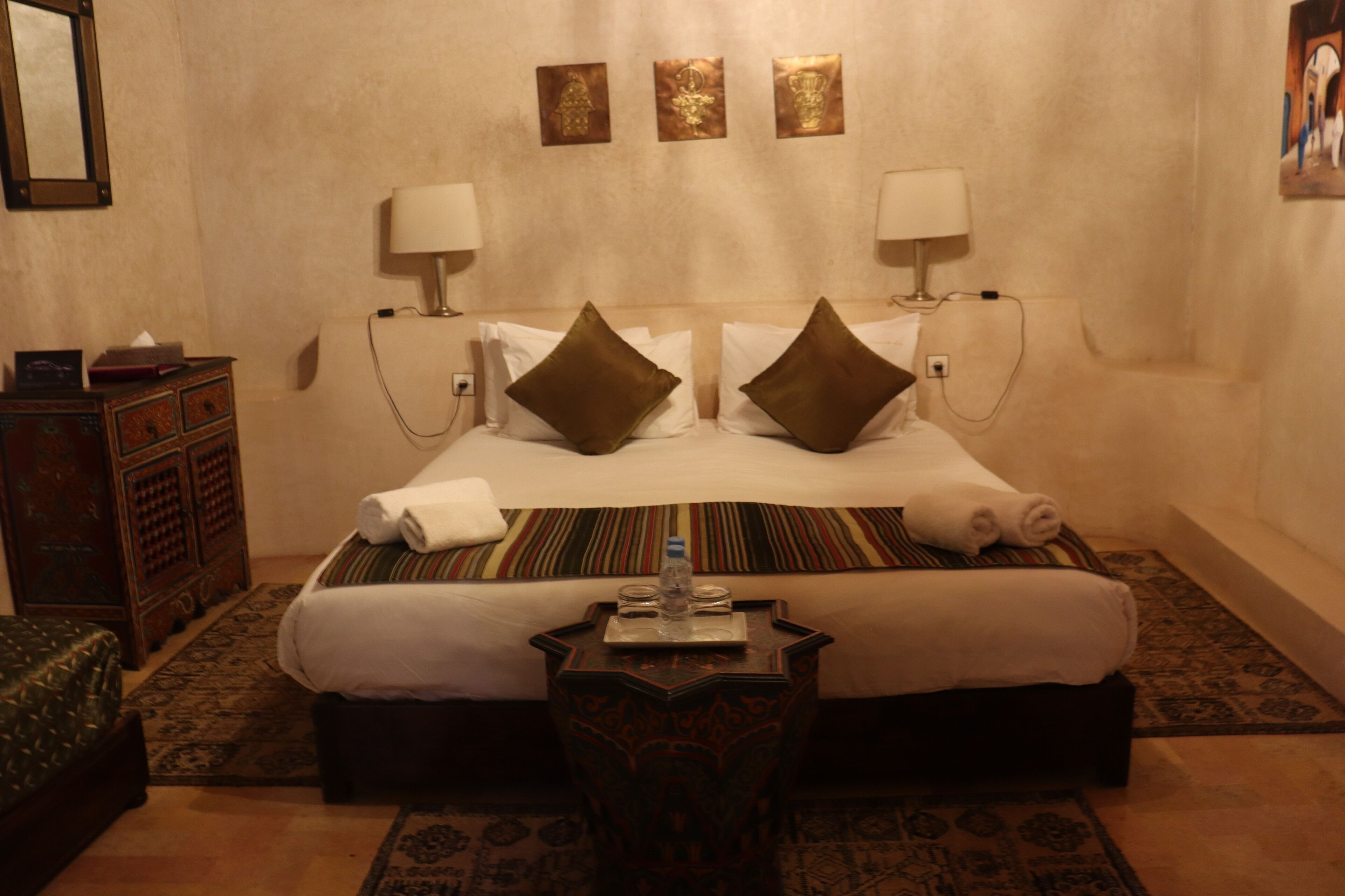 A hotel room with a large bed, two bedside lamps, a small table with a bottle and glasses, and a decorative mirror on the left.
