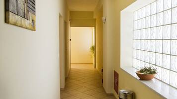 A hallway with a tiled floor, a trash can, a potted plant, and a window with a textured pattern.