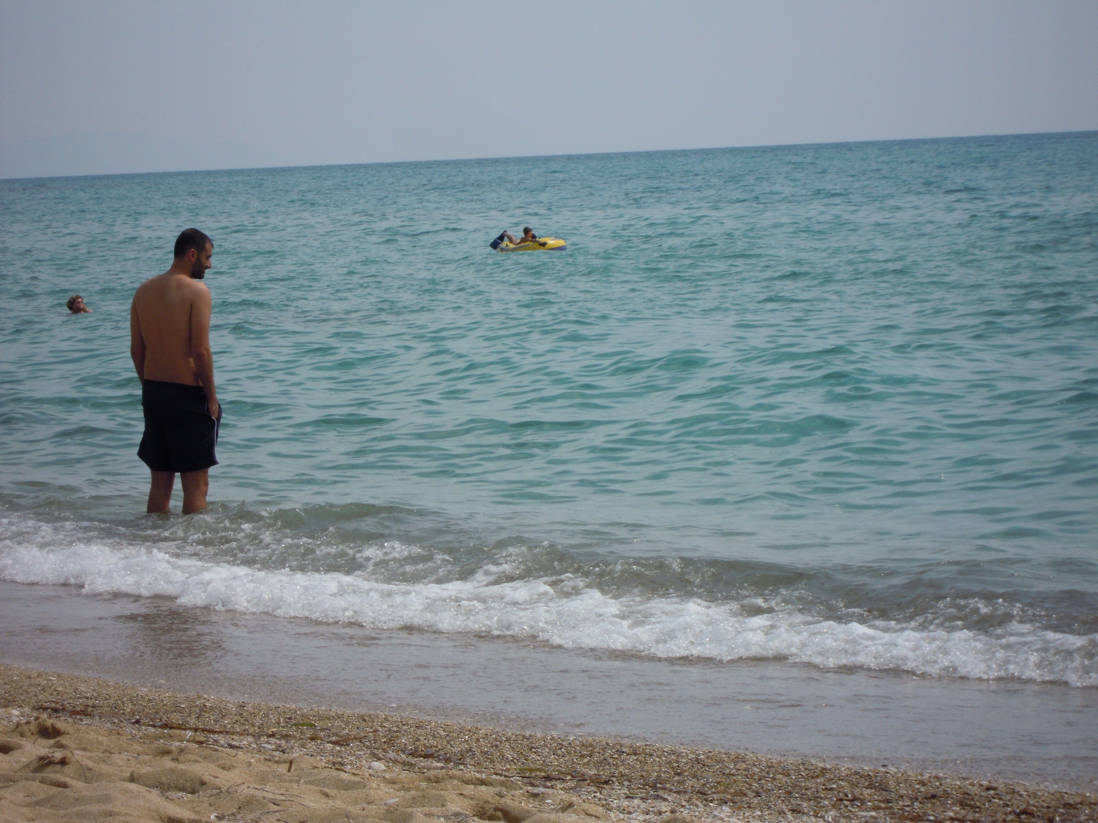 A beach scene with a man standing in shallow water, a yellow raft with two people in the distance, and another person swimming in the water.