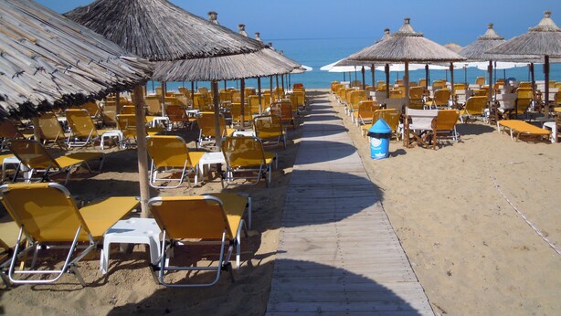 Un hotel frente a la playa con filas de sillas amarillas y sombrillas de paja, un camino de madera y una vista despejada del mar.