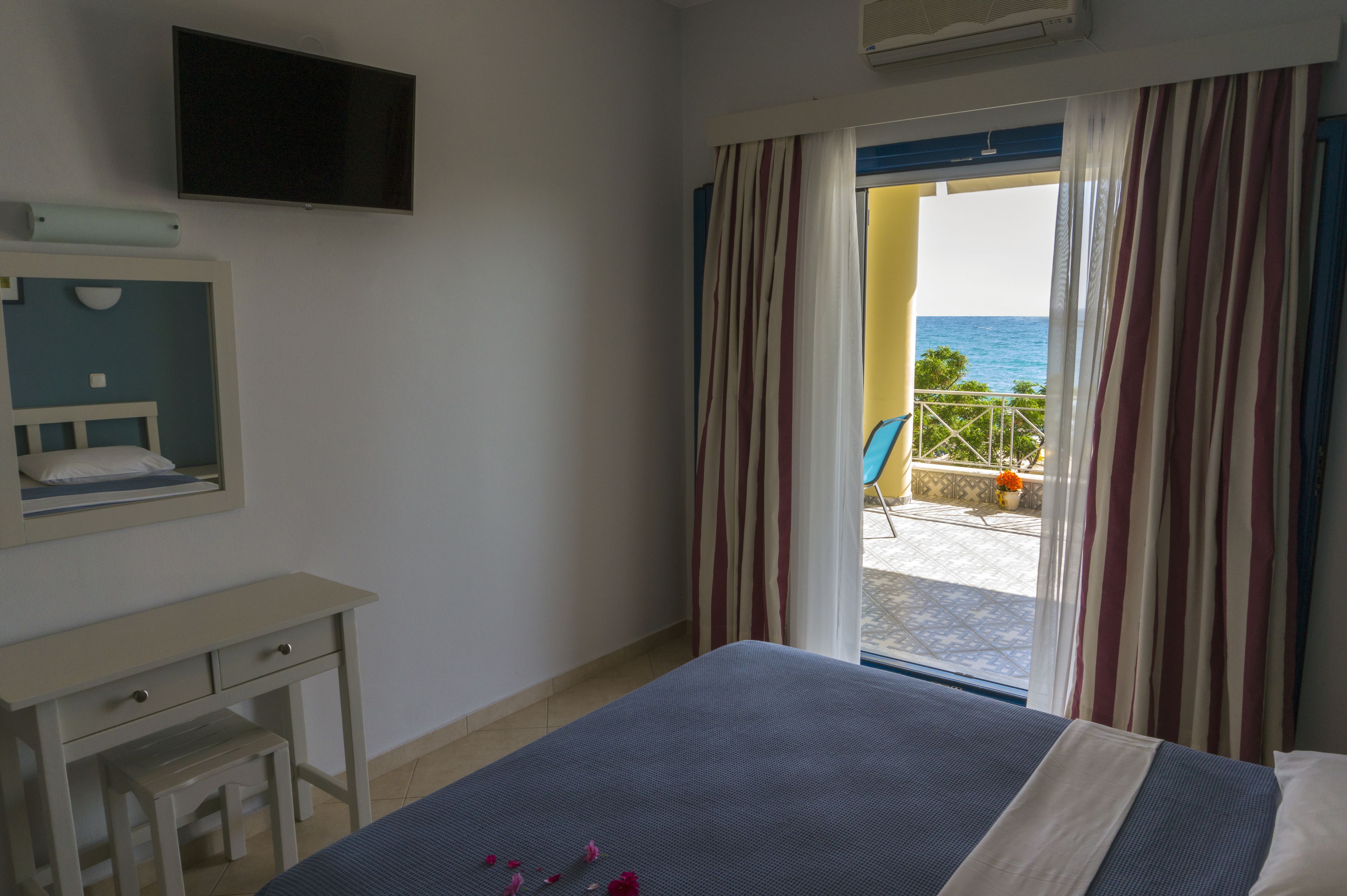 A hotel room with a bed, a desk, a mirror, and a TV. There is a balcony with a view of the sea.
