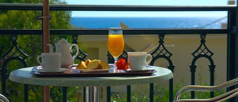 A round table with a tray of breakfast items, including bread, a glass of orange juice, and two cups, set against a balcony railing with a view of the sea.