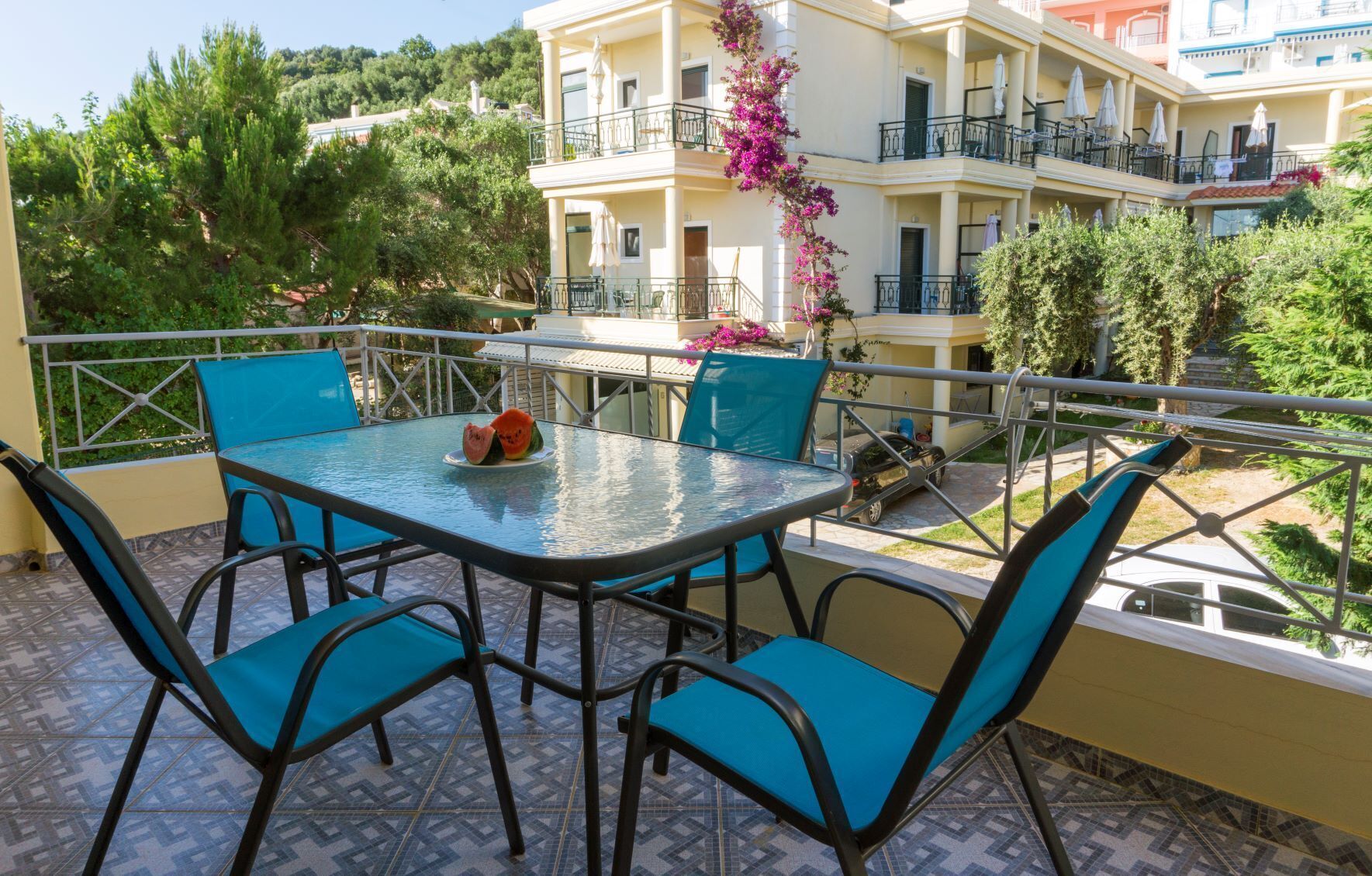 A balcony with a glass table and blue chairs, overlooking a building with balconies and a garden.