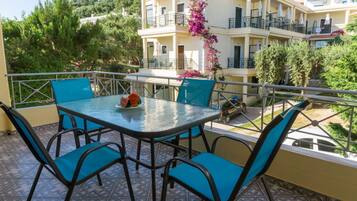 A balcony with a glass table and blue chairs, overlooking a building with balconies and a garden.