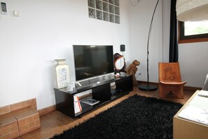 Uma sala de estar com uma TV de tela plana em um suporte preto, uma mesinha com um vaso e livros, um tapete preto e uma cadeira de madeira com padrão de mão.