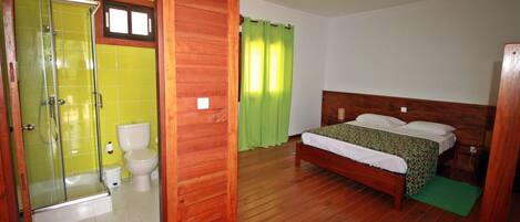 A room with a wooden floor, a bed with a green bedspread, a wooden headboard, a shower with a glass door, and a toilet.