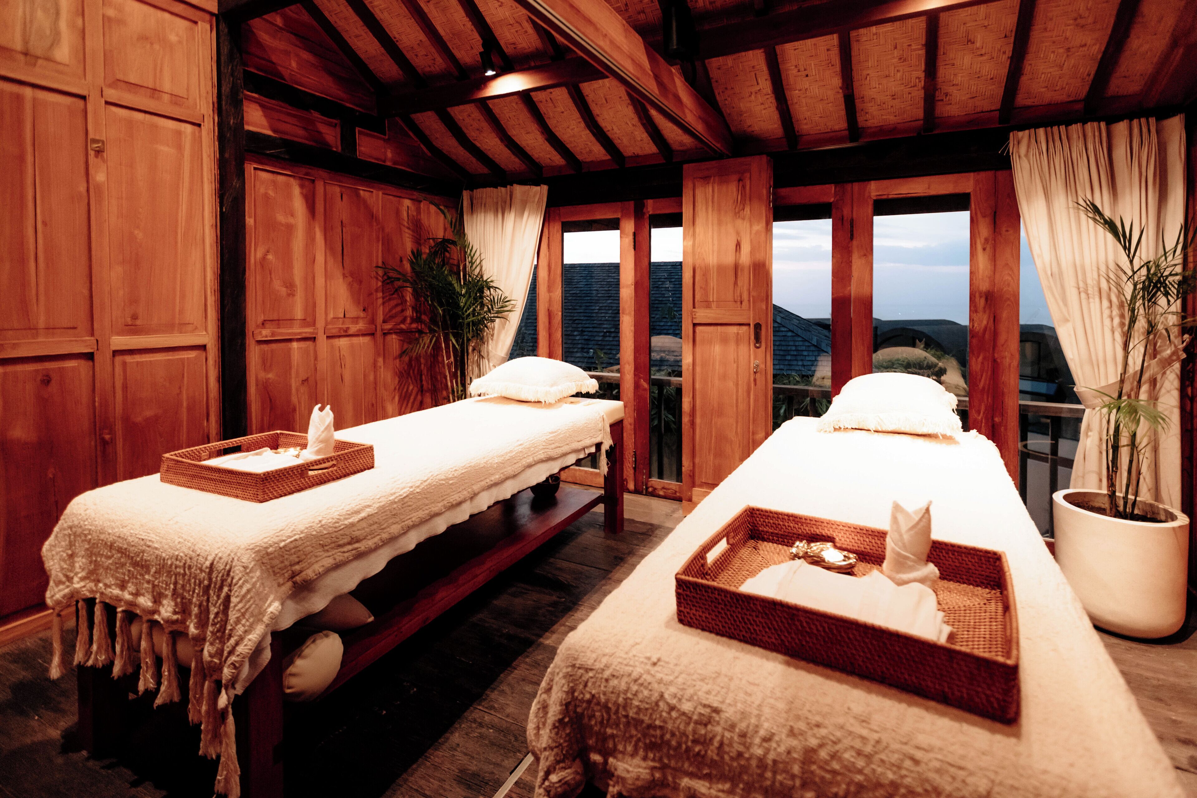 A room with wooden walls and a large window. There are two beds with white linens and baskets on them.