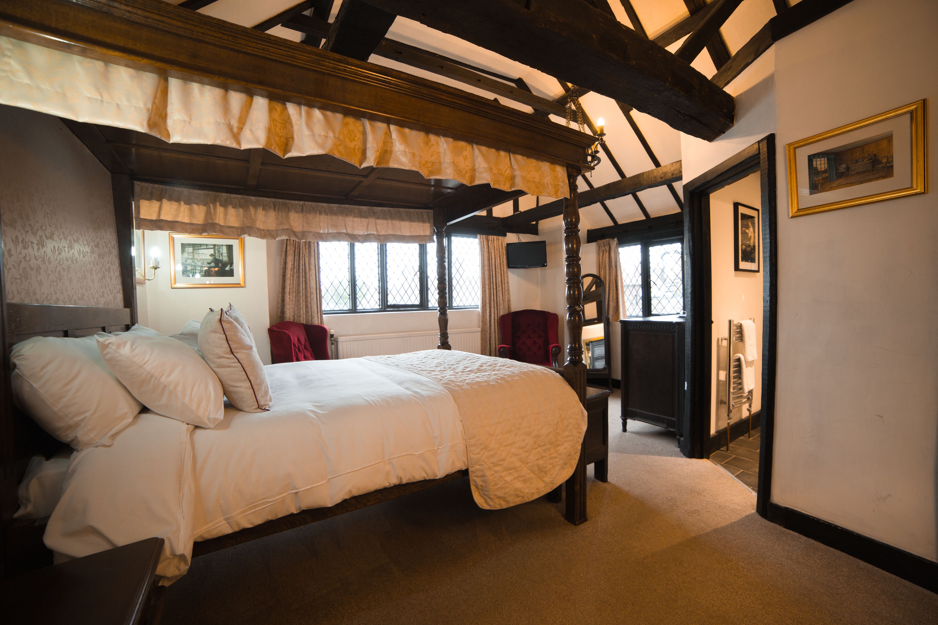 A traditional bedroom with a four-poster bed, a red armchair, and a wooden ceiling.