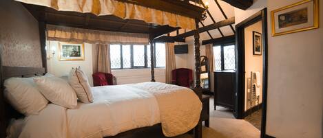 A traditional bedroom with a four-poster bed, a red armchair, and a wooden ceiling.