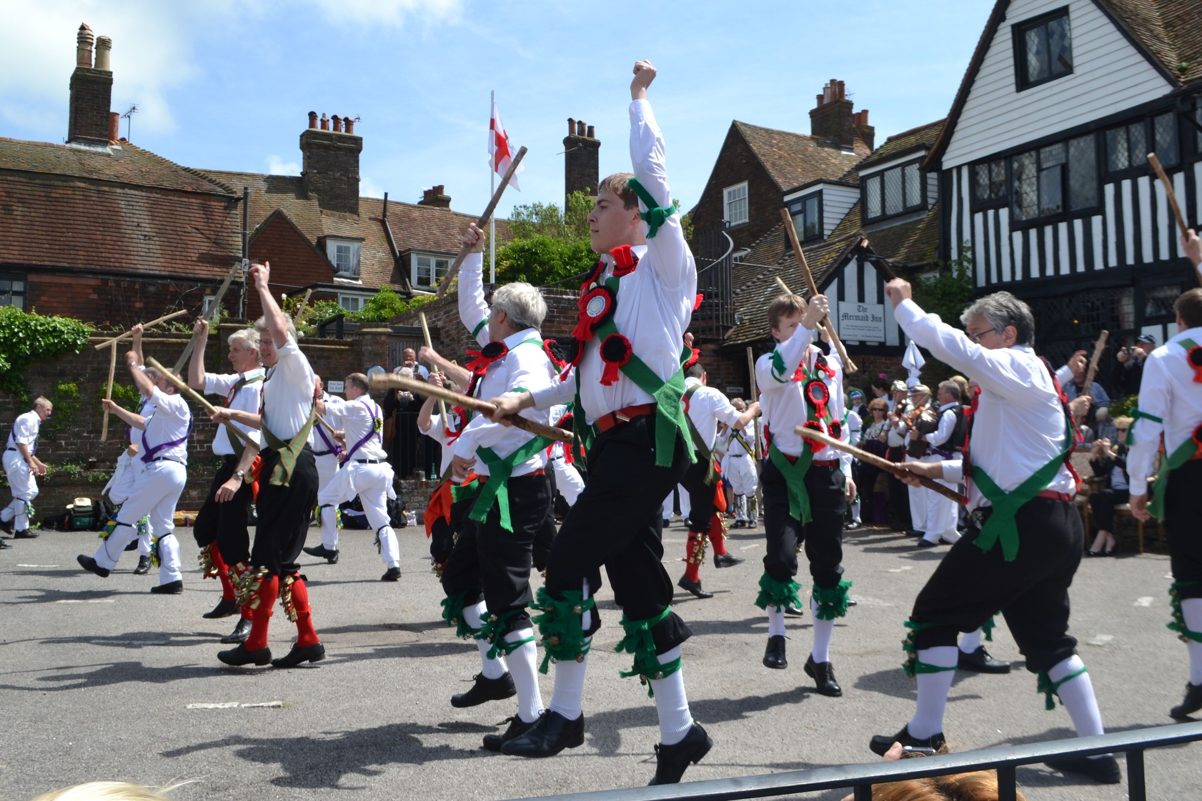 A group of people in traditional attire performing a dance in an outdoor setting with buildings in the background.