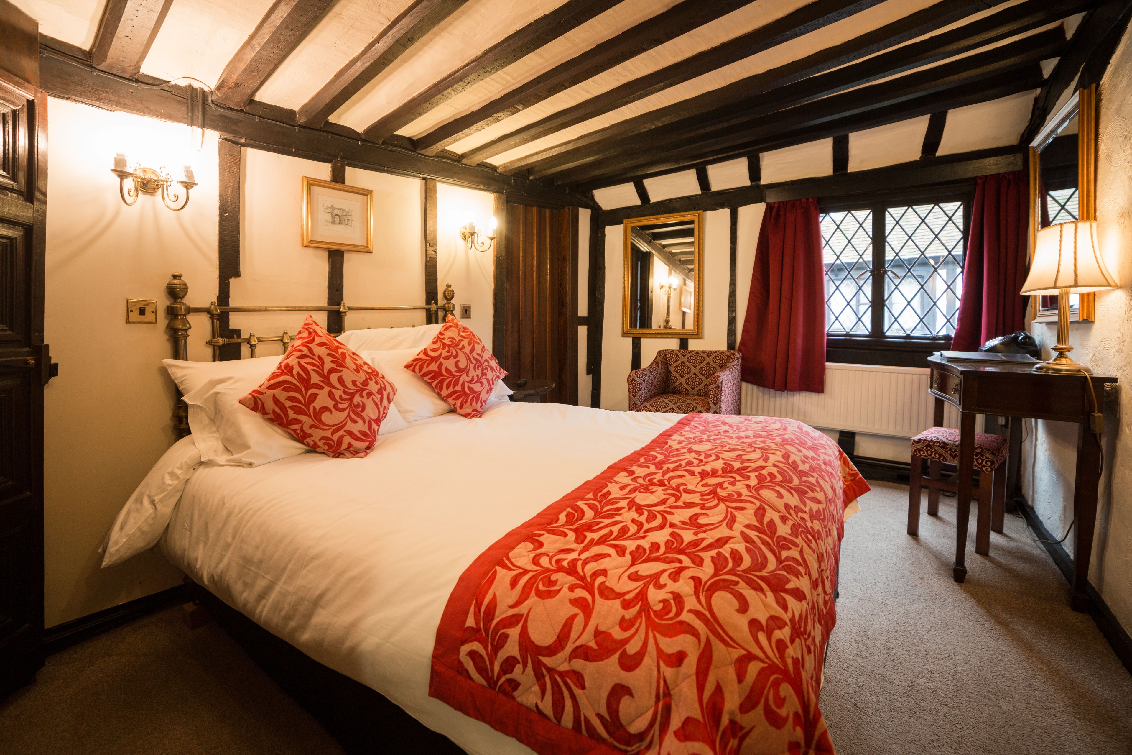 A traditional room with exposed wooden beams, a large bed with red and white bedding, a desk with a lamp, and a window with red curtains.