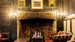 A traditional fireplace with a brick wall, a wooden mantel, and framed pictures above. There are leather sofas and a wooden table in the foreground. - The Mermaid Inn (Rye)