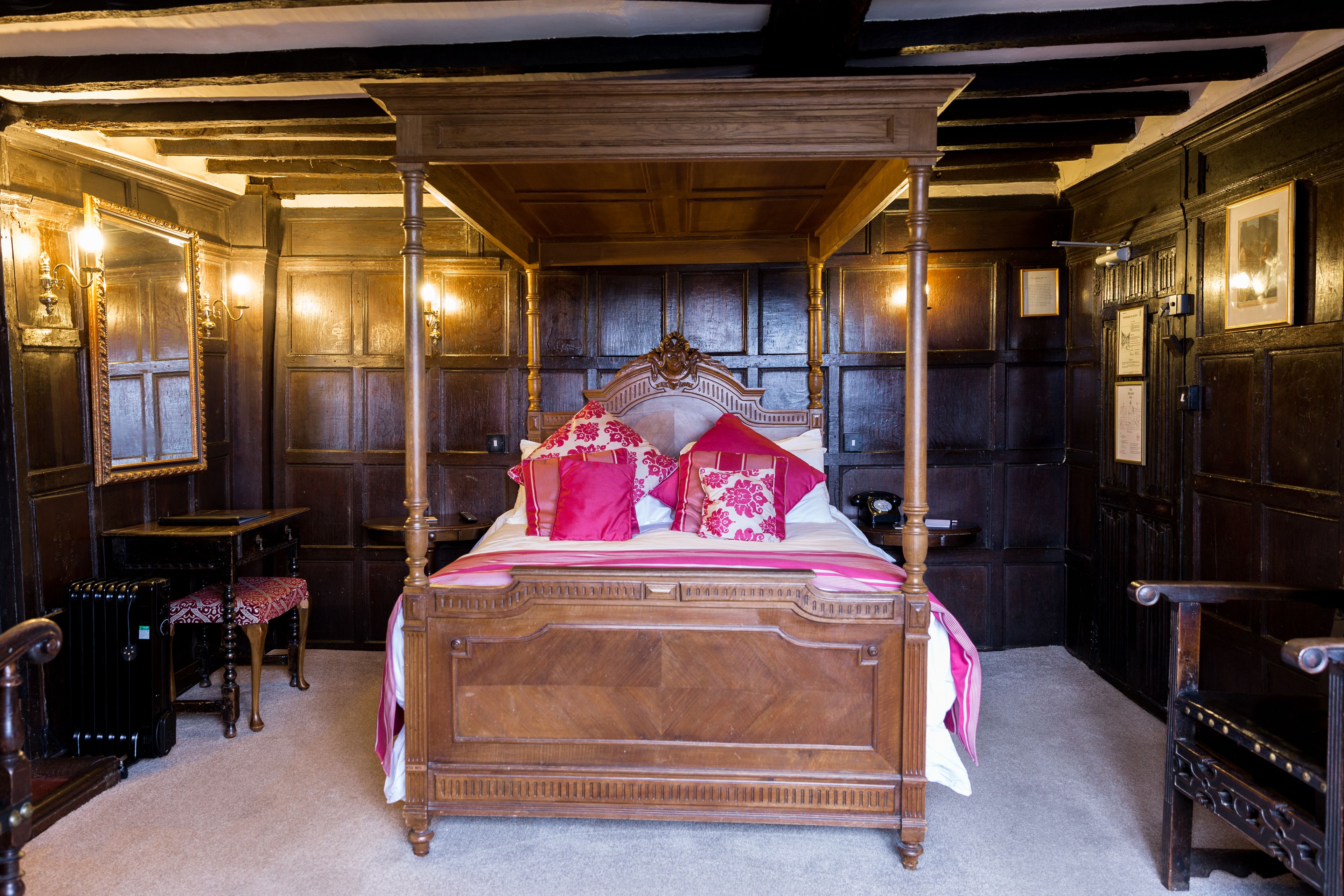 Un lit à baldaquin avec une tête de lit en bois, une couvre-lit rouge et blanche et des coussins assortis. Il y a deux tables de chevet avec des lampes et une chaise placée à gauche du lit.