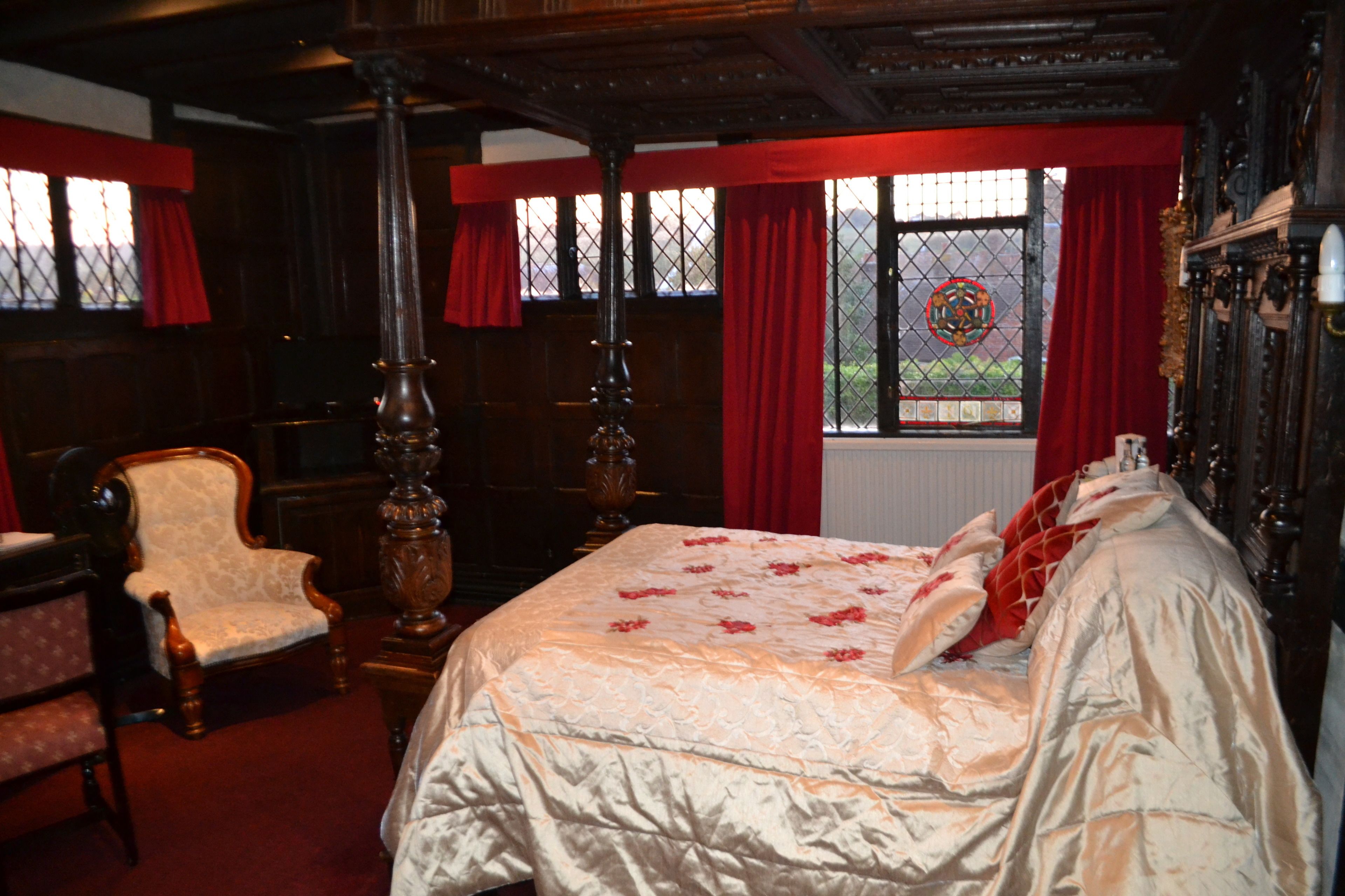 A traditional bedroom with a four-poster bed, a wooden chair, and a large window with a view of greenery.