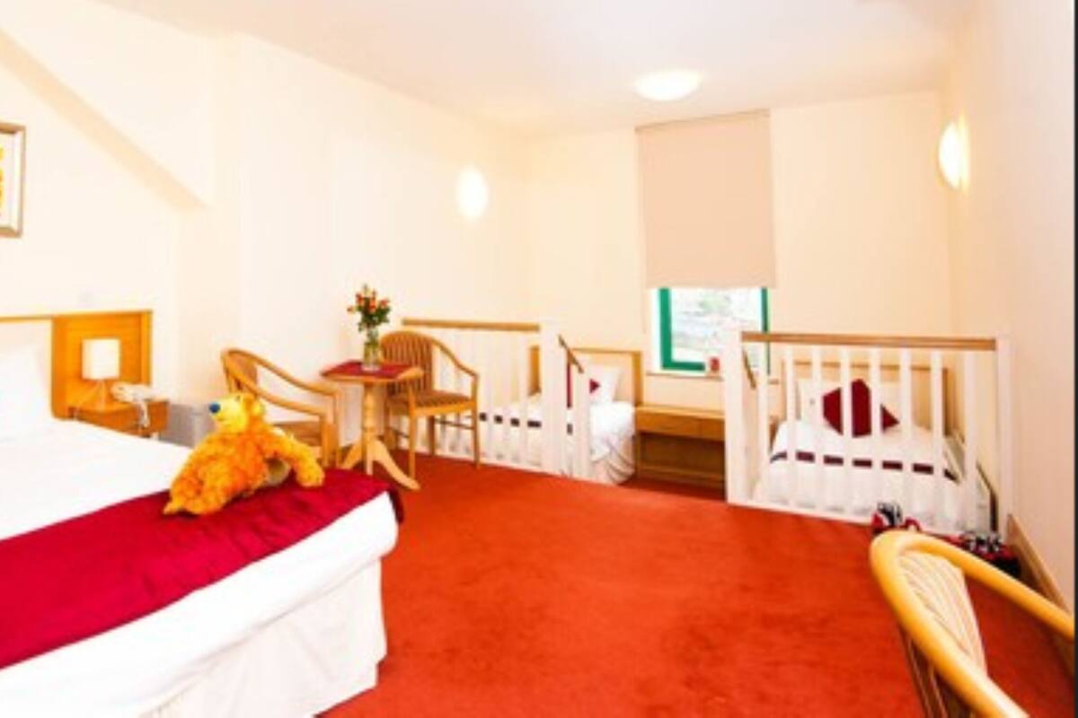 A hotel room with a bed, a red pillow, a teddy bear, a chair, a small table, and a crib.